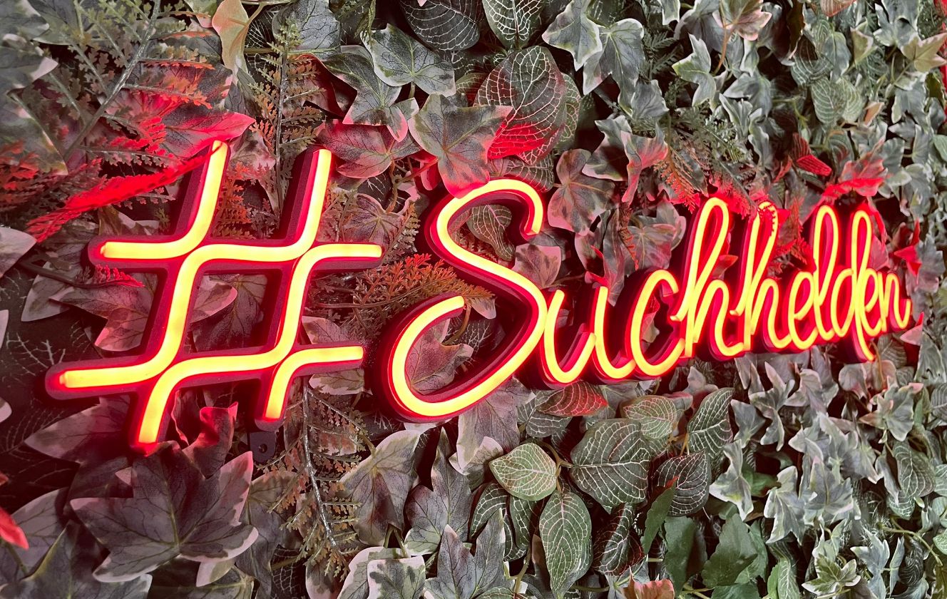 Leuchtendes Neon-Logo mit Schriftzug #Suchhelden vor grüner Blattwand