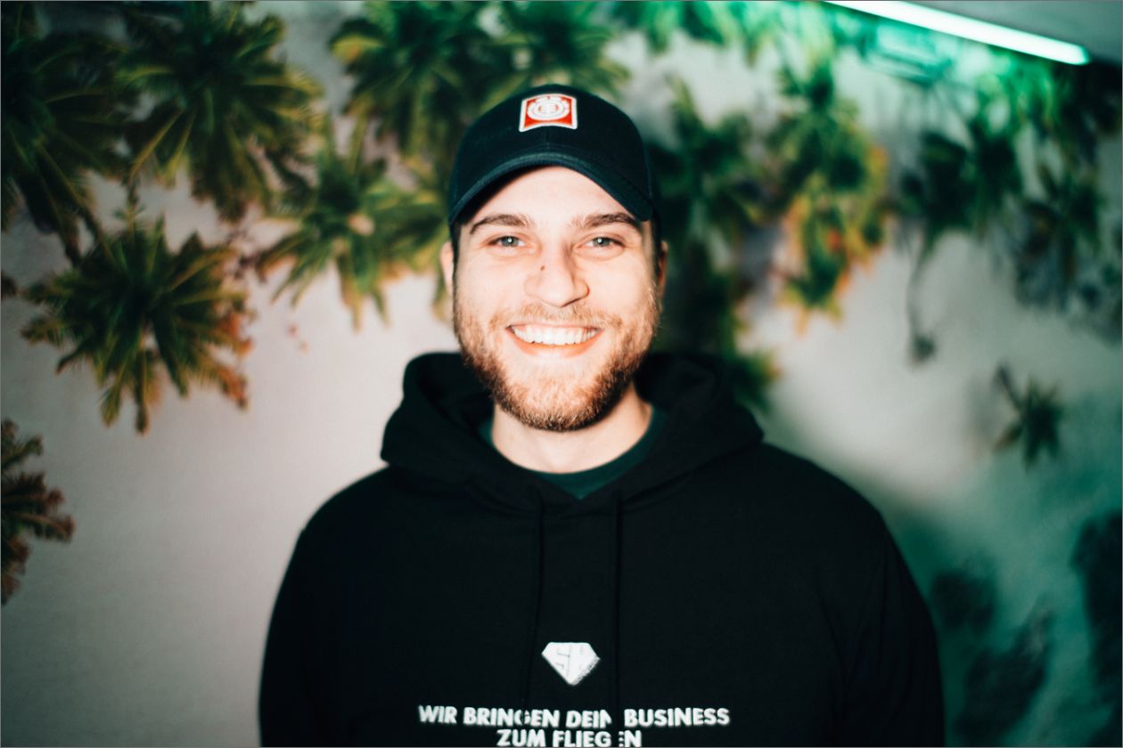 Malte Krekeler lächelt in schwarzem Hoodie vor Hintergrund mit Pflanzenmotiv.