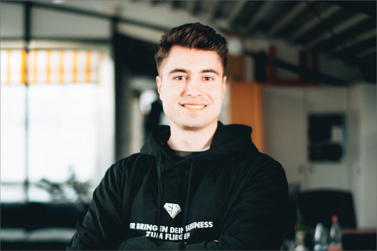 Malte Hundertmark lächelt, trägt schwarzen Hoodie steht in modernem Büro.